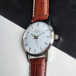 Op de afbeelding: Een zilveren Breitling Gen&egrave;ve polshorloge met een witte wijzerplaat en bruine leren band. Het horloge heeft een ronde wijzerplaat met zilveren wijzers en markeringen. De band is donkerbruin met witte stiksels.