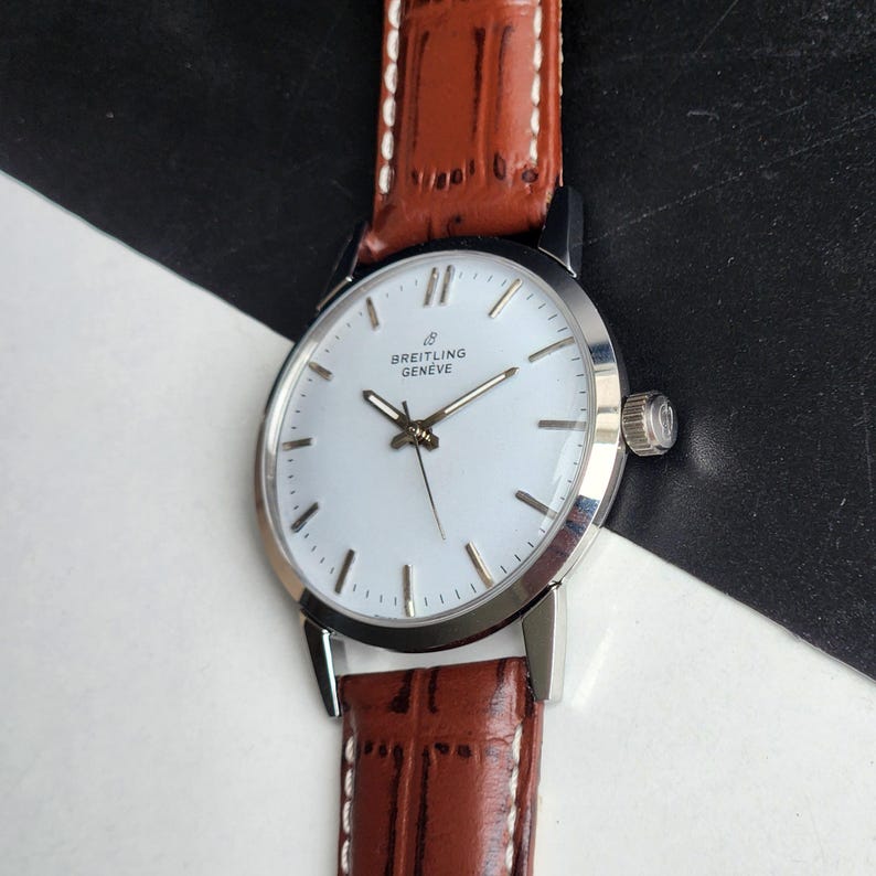 Op de afbeelding: Een zilveren Breitling Gen&egrave;ve polshorloge met een witte wijzerplaat en bruine leren band. Het horloge heeft een ronde wijzerplaat met zilveren wijzers en markeringen. De band is donkerbruin met witte stiksels.