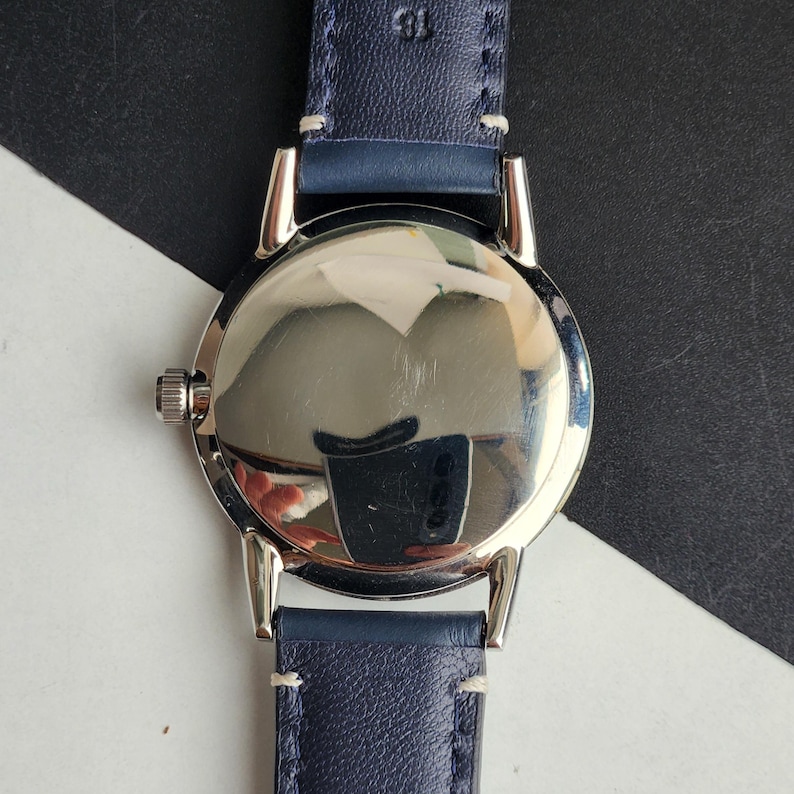 May include: A silver wristwatch with a navy blue leather strap. The watch face is round and reflective, with a small crown. The strap is stitched with white thread.