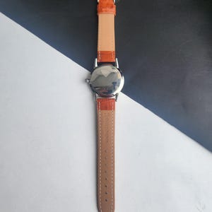 May include: A silver-toned wristwatch with an orange leather band. The watch face is reflective, and the band is a light brown color on the inside. The band is secured with a silver buckle. The watch is a classic design.
