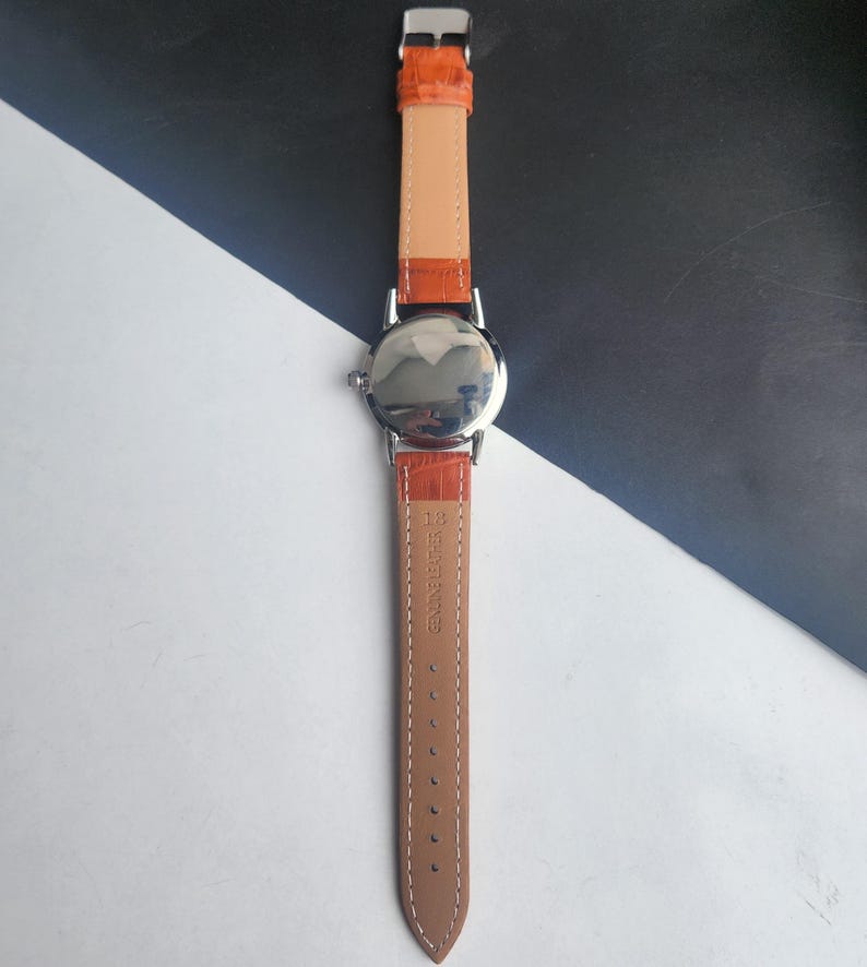 May include: A silver-toned wristwatch with an orange leather band. The watch face is reflective, and the band is a light brown color on the inside. The band is secured with a silver buckle. The watch is a classic design.