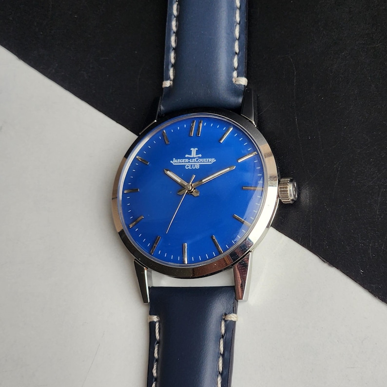May include: A silver wristwatch with a striking blue face and a blue leather strap. The watch face displays the text "Jaeger-LeCoultre CLUB". The strap is secured with white stitching.