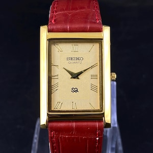 Op de afbeelding: Een goudkleurig Seiko Quartz horloge met een rechthoekige wijzerplaat en Romeinse cijfers. Het horloge heeft een crèmekleurige wijzerplaat, zwarte wijzers en een rode leren band. De wijzerplaat toont het Seiko-logo en het woord "QUARTZ".