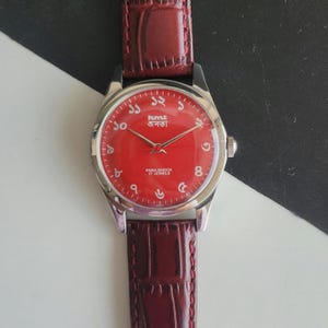 Puede incluir: Un reloj de pulsera con esfera roja y caja plateada. El reloj tiene una correa de cuero burdeos. La esfera muestra el texto "PuYLE জনতা" y "PARA SHOCK 17 JEWELS".