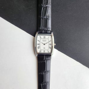 Könnte beinhalten: Eine rechteckige Seiko Quartz-Uhr mit weißem Zifferblatt, schwarzen römischen Ziffern und einem schwarzen Lederarmband. Die Uhr hat ein silberfarbenes Gehäuse und ein strukturiertes Armband. Die Uhr wird vor einem schwarz-weißen Hintergrund präsentiert.