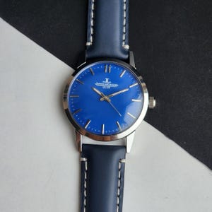 May include: A wristwatch with a blue face and a navy blue leather strap. The watch face has silver hands and hour markers. The watch has the text "JAEGER-LECOULTRE CLUB" printed on the face. The strap is stitched with white thread.