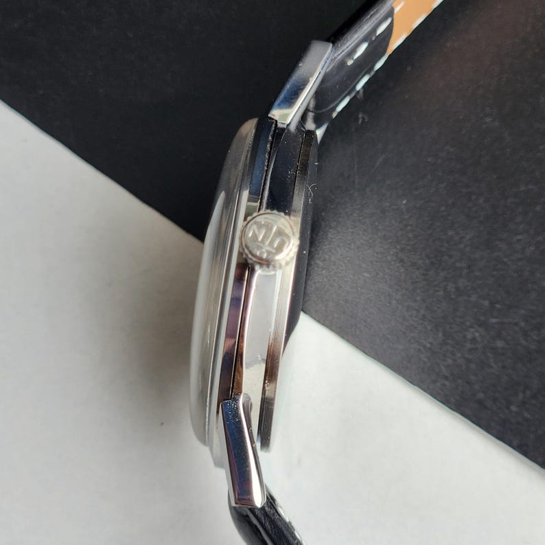 May include: A silver-toned wristwatch with a black leather band. The watch face is round with a silver bezel and a small crown. The band is stitched with white thread. The watch is a classic style.