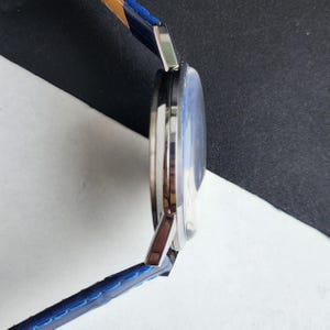 Puede incluir: Vista lateral de un reloj de pulsera plateado con esfera azul y correa de cuero azul. La correa tiene costuras azules y una parte inferior marr&oacute;n. El reloj est&aacute; sobre un fondo blanco y negro.