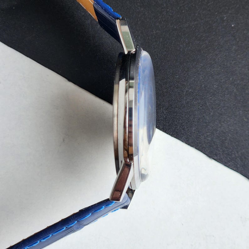 Puede incluir: Vista lateral de un reloj de pulsera plateado con esfera azul y correa de cuero azul. La correa tiene costuras azules y una parte inferior marr&oacute;n. El reloj est&aacute; sobre un fondo blanco y negro.