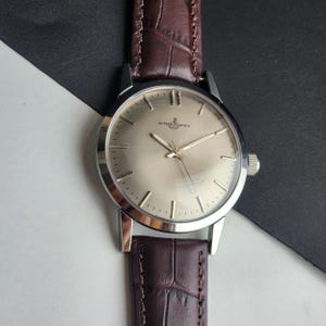 May include: A silver-toned wristwatch with a light gold face and a brown leather band. The watch face features the brand name "ULYSSE NARDIN" and a small anchor symbol. The band has a crocodile skin texture.