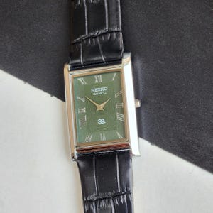 Pode incluir: Rel&oacute;gio Seiko Quartz retangular com mostrador verde e caixa prateada. O rel&oacute;gio tem algarismos romanos e uma pulseira de couro preta. O mostrador exibe o logotipo Seiko e a palavra "Quartz".