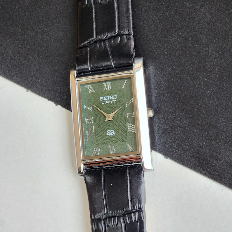 Pode incluir: Rel&oacute;gio Seiko Quartz retangular com mostrador verde e caixa prateada. O rel&oacute;gio tem algarismos romanos e uma pulseira de couro preta. O mostrador exibe o logotipo Seiko e a palavra "Quartz".