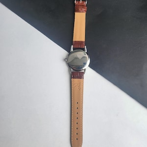 Op de afbeelding: Een zilverkleurig polshorloge met een bruine leren band. De wijzerplaat van het horloge is rond en reflecterend. De band is aan de binnenkant lichtbruin en aan de buitenkant donkerbruin. Het horloge wordt getoond tegen een zwart-witte achtergrond.