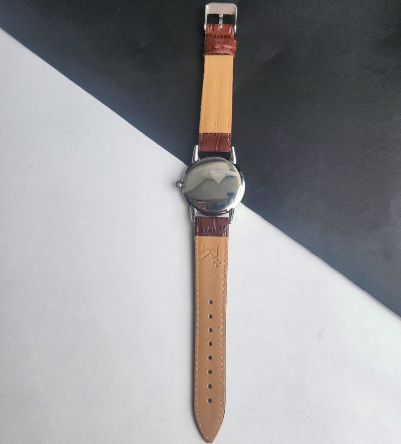 Op de afbeelding: Een zilverkleurig polshorloge met een bruine leren band. De wijzerplaat van het horloge is rond en reflecterend. De band is aan de binnenkant lichtbruin en aan de buitenkant donkerbruin. Het horloge wordt getoond tegen een zwart-witte achtergrond.