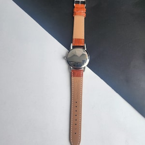 May include: A silver wristwatch with an orange leather strap. The watch features a round face and a light tan leather backing. The strap is fastened with a silver buckle. The watch is shown against a black and white background.