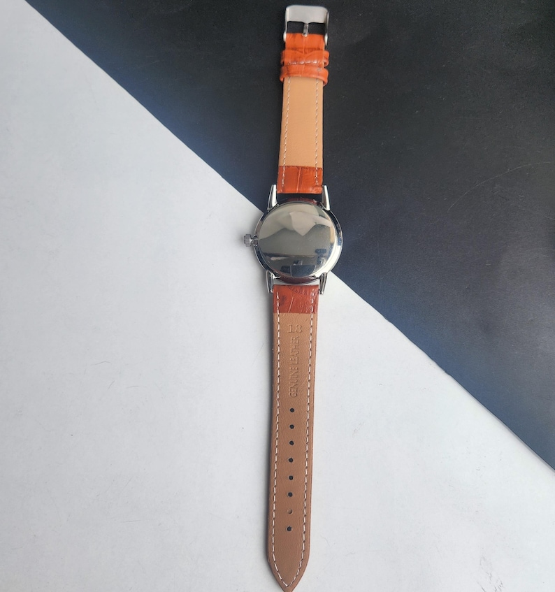 May include: A silver wristwatch with an orange leather strap. The watch features a round face and a light tan leather backing. The strap is fastened with a silver buckle. The watch is shown against a black and white background.