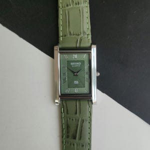 Peut inclure: Une montre Seiko Quartz rectangulaire avec un cadran vert et un bracelet en cuir vert assorti. Le cadran de la montre comporte des chiffres romains et le logo Seiko. Le bracelet a une texture peau de crocodile.