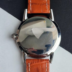 May include: Close-up of a silver-toned watch with a polished, reflective back. The watch has a brown leather strap with white stitching. The watch's design is simple and classic, with a focus on the materials and craftsmanship.