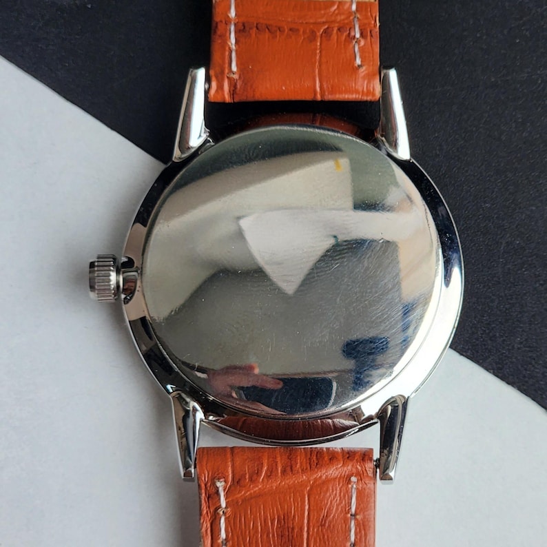 May include: Close-up of a silver-toned watch with a polished, reflective back. The watch has a brown leather strap with white stitching. The watch's design is simple and classic, with a focus on the materials and craftsmanship.