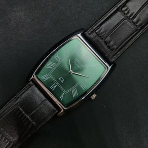 May include: A rectangular Seiko Quartz wristwatch featuring a sunray green dial with silver Roman numeral hour markers, silver hands, and a black leather strap with a textured crocodile-style pattern and a polished silver-tone metal buckle.