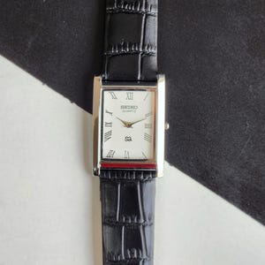 May include: A rectangular Seiko Quartz watch with a white face, Roman numeral hour markers, and a black leather strap. The watch has a silver-coloured case and a small seconds dial. The strap has a crocodile skin pattern.