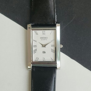 Może przedstawiać: Prostokątny zegarek Seiko Quartz z białą tarczą, rzymskimi cyframi i czarnym skórzanym paskiem. Zegarek ma srebrną kopertę i małe logo pod wskazówkami.
