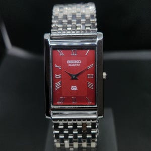 May include: A rectangular Seiko Quartz wristwatch with a vibrant red dial featuring a subtle wave pattern, silver-tone Roman numeral hour markers, and thin black hands. The watch has a polished silver-tone metal case and a matching link bracelet.