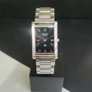 May include: A rectangular Seiko Quartz watch with a silver metal strap and case. The watch face is black with silver Roman numerals and hands. The watch is displayed on a black stand.