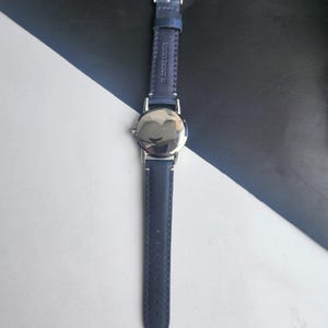 May include: A wristwatch with a navy blue leather strap and a silver watch face. The strap is stitched and has a silver buckle. The watch is shown against a black and white background.