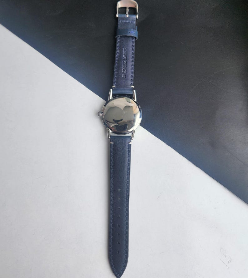 May include: A wristwatch with a navy blue leather strap and a silver watch face. The strap is stitched and has a silver buckle. The watch is shown against a black and white background.