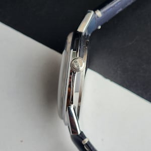 May include: A silver wristwatch with a dark blue leather strap. The watch face is round with a silver bezel. The watch has a silver crown and a dark blue leather strap.