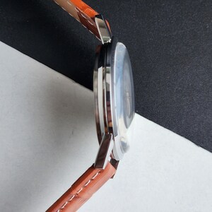 May include: A side view of a wristwatch with a brown leather band and a silver-colored case. The watch face is visible, with a light-colored dial. The band is stitched with white thread. The watch is set against a black and white background.