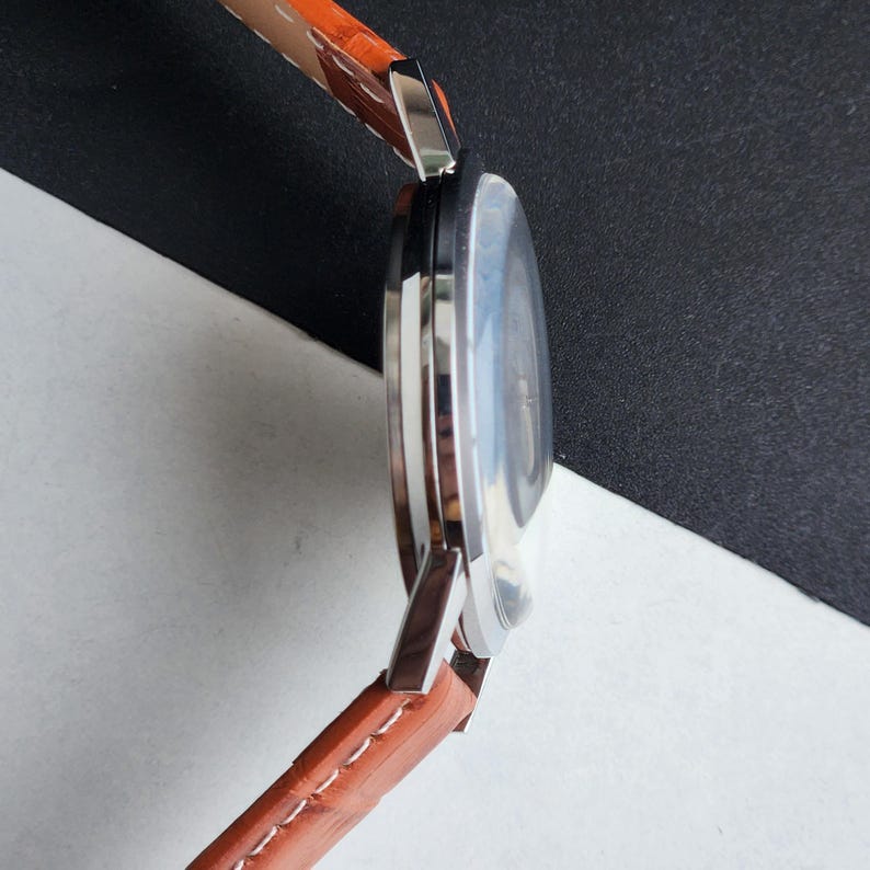 May include: A side view of a wristwatch with a brown leather band and a silver-colored case. The watch face is visible, with a light-colored dial. The band is stitched with white thread. The watch is set against a black and white background.