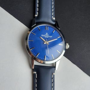 May include: A wristwatch with a blue face and silver case. The watch has a blue leather strap with white stitching. The watch face has silver hands and markers. The text "JAEGER-LECOULTRE CLUB" is printed on the watch face.