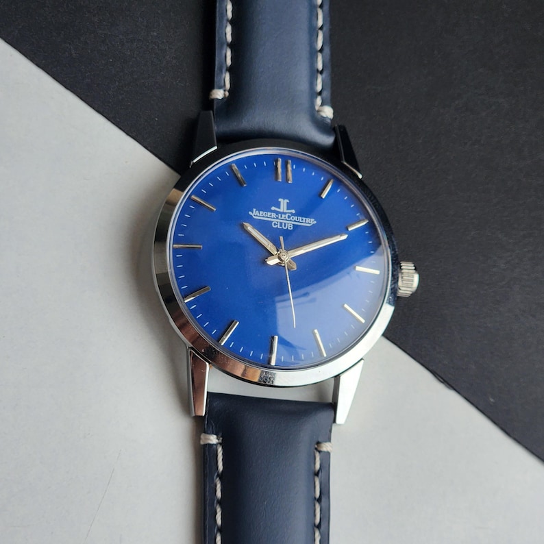May include: A wristwatch with a blue face and silver case. The watch has a blue leather strap with white stitching. The watch face has silver hands and markers. The text "JAEGER-LECOULTRE CLUB" is printed on the watch face.