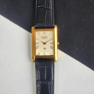 May include: A gold Seiko Quartz watch with a rectangular face and black leather strap. The watch face is cream-coloured with Roman numerals and gold hands. The strap has a textured, crocodile-like pattern.