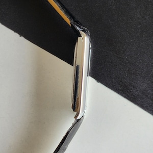 Pode incluir: Um rel&oacute;gio de pulso prateado com uma pulseira de couro preta. O mostrador do rel&oacute;gio &eacute; retangular e tem uma superf&iacute;cie polida e reflexiva. A pulseira &eacute; presa &agrave; caixa do rel&oacute;gio com um verso de couro amarelo-castanho.