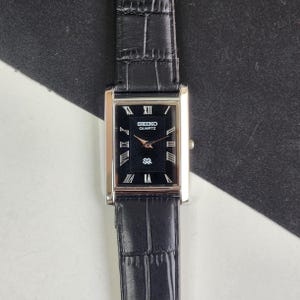 May include: A rectangular Seiko Quartz watch with a black face and silver-tone case. The watch features Roman numeral hour markers and a black leather band. The watch face displays the Seiko Quartz logo.