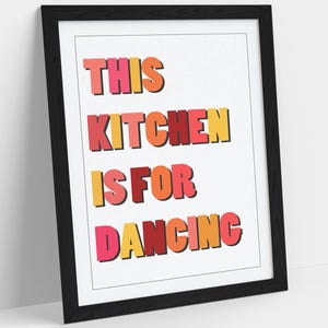 May include: A framed art print with the text "THIS KITCHEN IS FOR DANCING" in colorful, bold letters. The text is in shades of pink, orange, and yellow with a black outline. The frame is black.