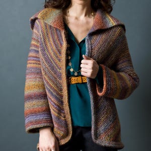 May include: A hooded, multi-colored knit jacket in shades of purple, orange, and brown. The jacket has a shawl collar and long sleeves. The model is wearing a teal blouse and a gold necklace. A brown belt cinches the waist.