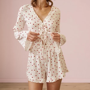 May include: White romper with red heart pattern. The top has a V-neck, long sleeves, and a lace-up front. The shorts have a ruffled hem. The outfit is made of a lightweight fabric.