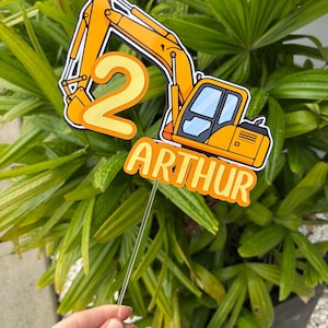 Könnte beinhalten: Ein Tortenstecker mit einer gelben Baustellenbagger-Illustration, der Zahl 2 und dem Namen Arthur aus orangefarbenem und gelbem Schichtkarton, befestigt an einem transparenten Kunststoffstab zur Dekoration von Torten oder Desserts.
