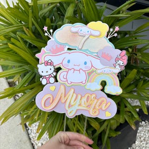 Puede incluir: Adorno para tarta multicapa con personajes de dibujos animados, incluyendo Hello Kitty y Cinnamoroll, con un arcoíris y el nombre "Myora" en cursiva. El adorno es principalmente de colores pastel con detalles brillantes.