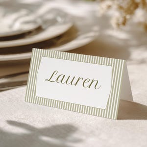 Puede incluir: Una tarjeta de sitio de papel plegada que presenta un panel central blanco con el nombre Lauren impreso en una fuente cursiva de color verde oliva oscuro, rodeado por un borde de rayas verticales de color verde oliva y blanco sobre cartulina texturizada.