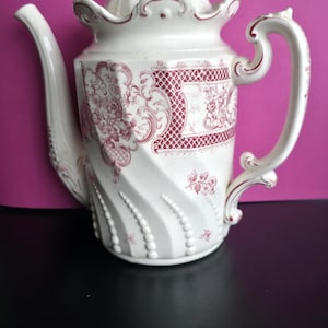 Op de afbeelding: Witte keramische theepot met deksel, tuit en handvat. De theepot heeft een rood bloemmotief en een gestructureerd, geribbeld ontwerp. Het handvat heeft een decoratieve, gebogen vorm. De theepot staat op een zwart oppervlak tegen een roze achtergrond.