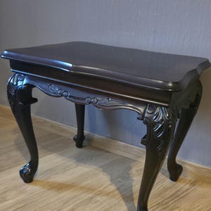 Peut inclure: Une table d'appoint en bois brun foncé dotée d'un plateau rectangulaire lisse aux bords incurvés et de quatre pieds galbés. La ceinture de la table est ornée de motifs sculptés en forme de feuilles qui descendent sur le haut de chaque pied.