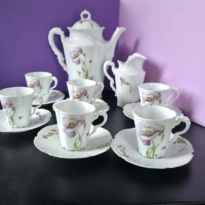 Puede incluir: Un juego de té de porcelana blanca con un delicado patrón de flores de amapola moradas. El conjunto incluye una tetera alta, una jarra para crema y cuatro tazas a juego con platillos. Cada pieza tiene un borde festoneado y acabado brillante.