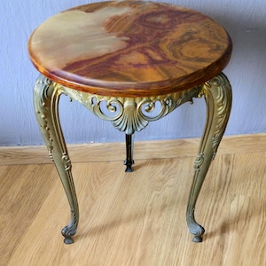 May include: A round side table featuring a polished, multi-toned brown and cream stone top with natural veining. The table is supported by three ornate, curved brass-colored metal legs with intricate scrollwork and shell-like detailing.