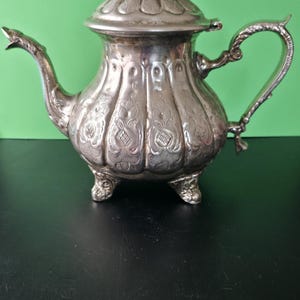 Op de afbeelding: Een zilverkleurige metalen theepot met een sierlijk, in reliëf aangebracht bloemmotief over het bolvormige lichaam. De pot heeft een gebogen tuit, een decoratief handvat, een deksel met een kleine knop en staat op drie korte, gedetailleerde voetjes.
