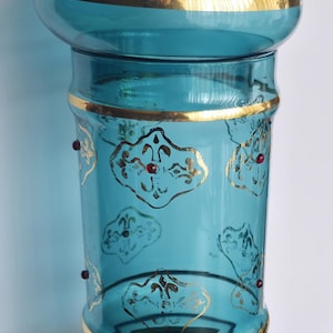 Peut inclure: Un vase en verre turquoise avec un dessus noir et une bordure dorée. Le vase est orné de motifs de filigrane dorés et de petits accents rouges. Le vase mesure environ 25 cm de haut.